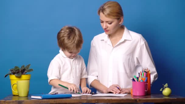 Molto bene. Affascinante madre sorridente seduta a tavola e che insegna al suo piccolo figlio a disegnare. Madre e figlio disegnano isolati su sfondo blu. Concetto per le giovani generazioni. Madre e figlio . — Video Stock