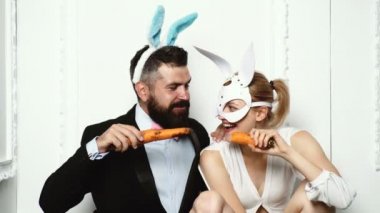 Tavşan kulaklı takım elbiseli beyefendi ve yüzünde deri maskeli bir sarışın havuç yiyor. Tavşan kulaklı komik bir çift. Havuç yemek gibi. Tüm aile için vitaminler. Beyaz üzerine izole.