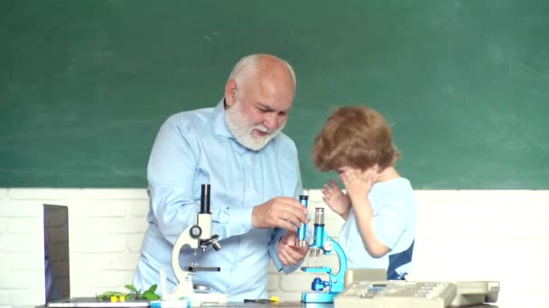 Счастливый милый умный мальчик и старый репетитор с микроскопом. Забавный маленький ребенок веселится на фоне школьной доски. Дедушка и ребенок учатся в классе . — стоковое видео