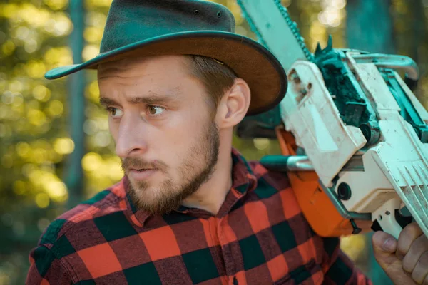 Lumberjack - retrato de cerca. Un leñador con motosierra en las manos. Cortador de madera con hacha o motosierra en el bosque de verano. Trabajador de madera caminando en el bosque con motosierra . —  Fotos de Stock