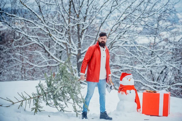 Lustiger Weihnachtsmann posiert mit Axt und Weihnachtsbaum. Ein Mann mit Bart trägt einen Weihnachtsbaum. Holzfäller mit Weihnachtsbaum im Winterpark. — Stockfoto