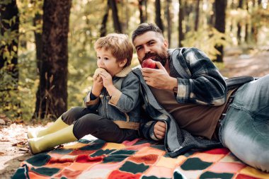 Sağlıklı yaşam tarzı. Doğa pikniği. Sağlıklı atıştırmalık. Vitamin yükü. Aile pikniği. Hipster sakallı baba oğluyla birlikte ormanda vakit geçiriyor. Acımasız sakallı adam ve küçük çocuk elma yiyor. Orman piknik yürüyüş