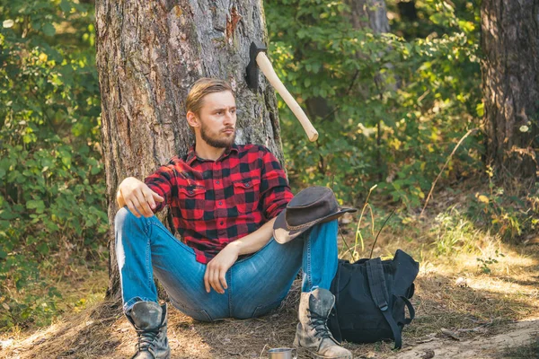 Trabalhador lenhador sentado na floresta. Homem bonito lenhador com um machado grande examina a árvore antes de cair . — Fotografia de Stock
