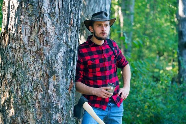 Skogshuggare med yxa på skogs bakgrund. Man gör mans jobb. En stilig ung man med skägg bär ett träd. Snygg ung man poserar som skogshuggare. — Stockfoto