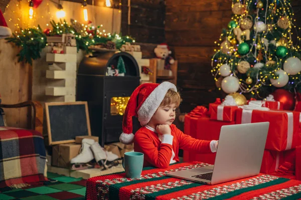 Vánoční nákupy. Služby dárků. Malý génius. Santa malý pomocník. Malý chlapeček z klobouku a kostýmů zábavy. Dítě s laptopem v blízkosti vánočního stromu. Kupte si vánoční dárky online — Stock fotografie