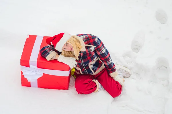 白い雪の背景に立って、両手で巨大な赤いギフトボックスを保持している幸せな女性。大きな贈り物を持つクリスマスの女の子。クリスマスガールプッシュ大きなギフトボックスフルレングスコンセプト. — ストック写真