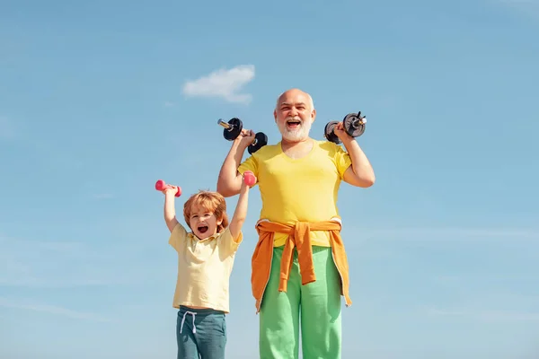 Nagyapa segítő gyerek a súlyzók. Mosolygós kisfiú és boldog csinos öreg ember gyakorolja a súlyzók. Egészséges neveltetés. — Stock Fotó