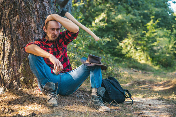 Lumberjack man sitting in the forest. Illegal logging continues today. Lumberjack worker standing in the forest with axe. Agriculture and forestry theme. Lumberjack standing with axe on forest.