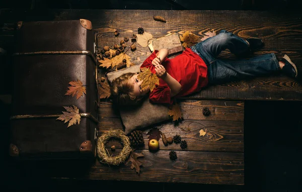 Little child boy lies on a warm blanket dreams of warm autumn. Blonde little boy resting with leaf on stomach lies on wooden floor in autumn leaves. Cute little child boy are getting ready for autumn.