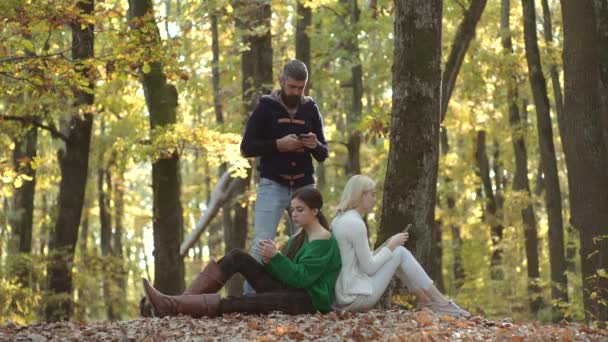 Redes sociales. Estudiantes en línea y e-learning outdoor. Grupo de dos chicas con un hombre guapo sosteniendo el teléfono móvil en las manos. Mujer escribiendo escribir mensaje en el teléfono inteligente en el parque de otoño . — Vídeos de Stock