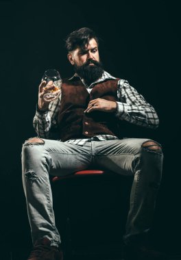 Takım elbiseli sakallı ve bıyıklı hipster, iş gününden sonra alkol içer. Alkol bağımlısı. Sakallı adam brendi bardağı tutar.