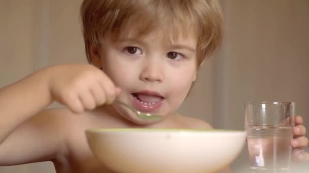 幸せな男の子のスプーンは自分自身を食べます。幸せな子供は朝食を食べます。幸せな子供だハイチェアに座ってキッチンの背景に食べるかわいい子供の男の子の男の子を笑う. — ストック動画