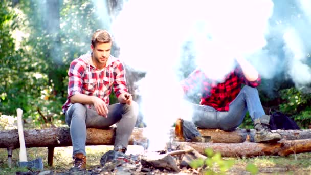 Dois homens bonitos amigos no acampamento perto da fogueira. Turistas relaxantes. Dois amigos lenhadores sentados na floresta. Amigos gostam de acampamento de fim de semana na floresta. Amigos se divertindo no acampamento fogo . — Vídeo de Stock