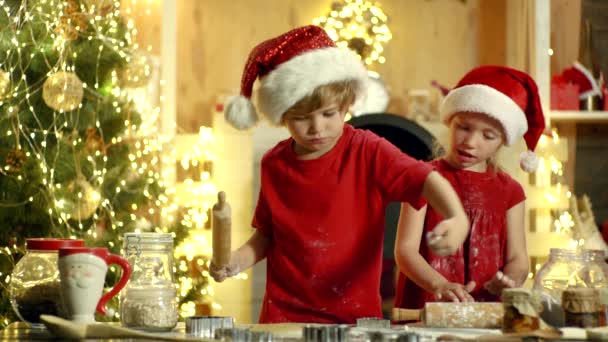 Kucharz świąteczny - dzieci w czapce Mikołaja. Dwoje dzieci zrobić piernik ciasteczko dla Mikołaja na tle światła Bożego Narodzenia. Mleko i ciasteczka dla Świętego Mikołaja. Kuchnia świąteczna. — Wideo stockowe