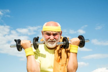 Dambıl kaldırarak egzersiz yapan mutlu bir son sınıf öğrencisi. Mavi gökyüzü arka planında büyük baba sportmen portresi. Spor merkezinde kıdemli sporcu..