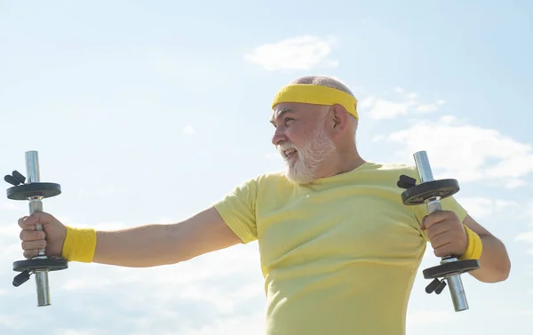 Sportivo senior nel centro sportivo. Sollevo manubri. Cura del corpo e assistenza sanitaria. Uomo anziano che si allena sul cielo blu. Salute stile di vita allegro . — Foto Stock