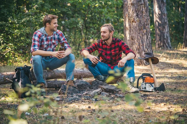Gruppe von zwei männlichen Rucksacktouristen, die sich am Lagerfeuer entspannen. junger Mann beim Picknick im Wald. Freunde sitzen beim Waldpicknick. glückliche junge Freunde beim Picknick auf dem Land. — Stockfoto