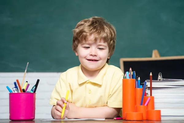 Little boy pupil with happy face expression near desk with school supplies - school concept. Funny little kid pointing up on blackboard. Kids with a book.