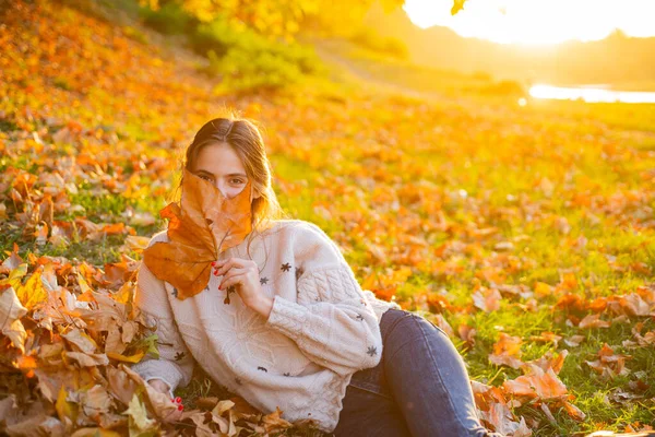 Jesienny flirt blondynka marzycielski pozowanie w listopadowym dniu. Zewnętrzne zdjęcie zadowolonej, długowłosej kobiety siedzącej na ziemi i trzymającej liść sykamora na tle liści. Piękno oczu. — Zdjęcie stockowe