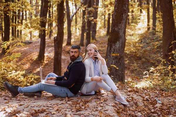 Pareja de negocios en otoño a pie. Pareja de negocios - hombre navegando por Internet en un portátil, mujer usando el teléfono móvil al aire libre. Negocios al aire libre en otoño parque . — Foto de Stock