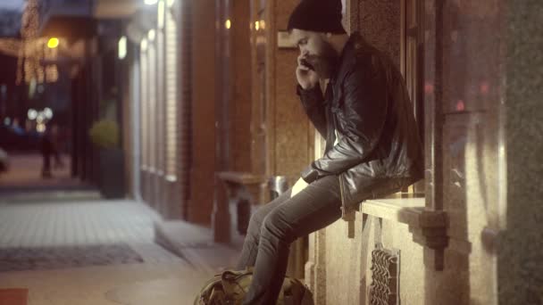 Bonito jovem hipster com seu smartphone lá fora na cidade noturna. Um homem bonito nas luzes da cidade à noite . — Vídeo de Stock