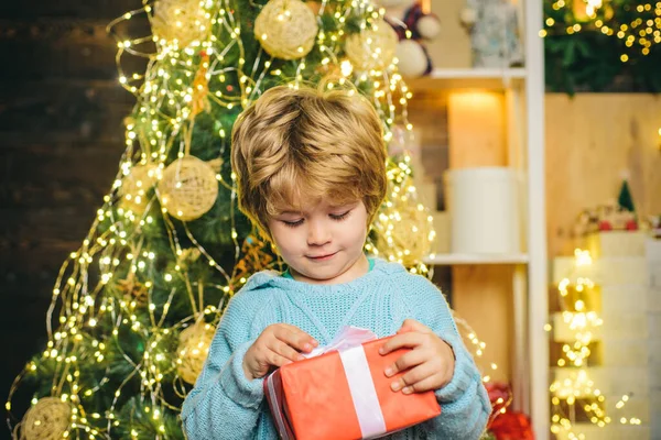 Happy child with Christmas gift. Portrait of Santa kid with gift looking at camera. Kid having fun near Christmas tree indoors. Happy child holding a red gift box with both hands.