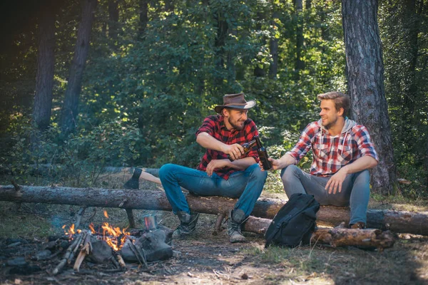 Two handsome men friends on camping near campfire. Tourists relaxing. Two friends lumberjack worker sitting in the forest with beer. Friends enjoy weekend camp in forest.