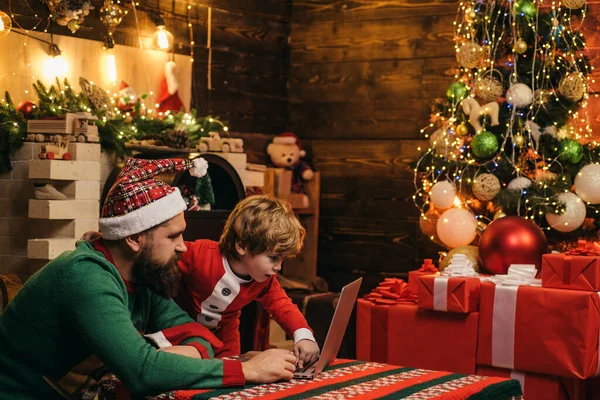 Social media. Happy little child son with father dressed in winter clothing think about Santa using computer near Christmas tree. New year Christmas concept. Gift emotions.