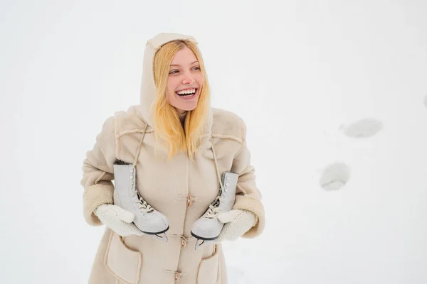 Ung blond kvinna i tröja och rolig hatt håller vita skridskor över axeln i iskalla vinterdag. Vacker kvinna promenader i vinter park och känsla underbart. — Stockfoto