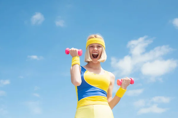 Eccitato giovane sportiva attraente indossa giallo con uniforme blu di formazione dimostrando esercizi con manubri rosa su sfondo cielo con nuvole. Moda e tendenze nello sport. Salute e sicurezza — Foto Stock