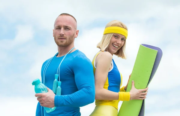 Couple heureux faisant de l'exercice à l'extérieur dans la nature. Fermez les athlètes à l'entraînement matinal. Fort bel homme faisant des exercices d'haltère debout à côté de la jeune athlète femelle avec tapis de yoga et bouteille de — Photo