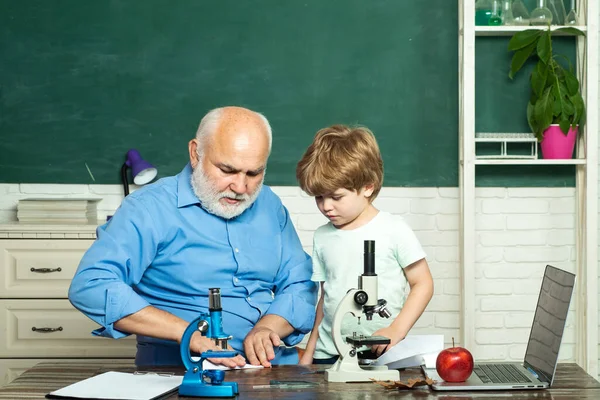 Un poco listo para estudiar. Padre e hijo. Día del maestro. Feliz chico inteligente lindo y viejo tutor con microscopio . — Foto de Stock