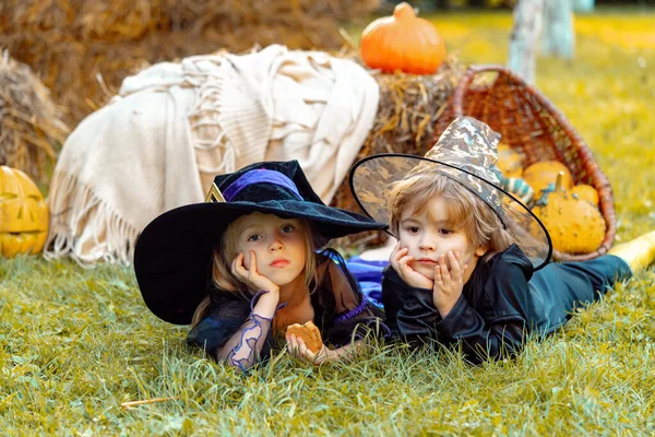 Gyerekek húga és testvére tökkel öltözve, csontváznak és boszorkánynak öltözve a Halloween partira. Boldog Halloween Gyermek lány és fiú ül széna vagy szalma a réten ősszel. — Stock Fotó