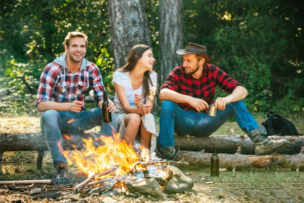 Enjoying camping holiday in countryside. Happy people sitting around campfire. Friends enjoy weekend barbecue in forest. Young people enjoying picnic in park on summer day.