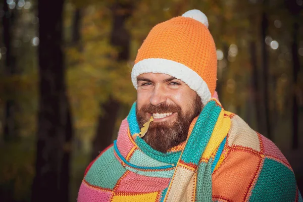 Wesoły hipster z brodą owinięty w wielokolorowy koc, w ciepłym kapeluszu, uśmiechnięty do kamery. Koncepcja odporności na choroby sezonowe, przeziębienia, zapobieganie katar. — Zdjęcie stockowe