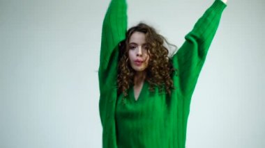 Kış kıyafetleri içinde, beyaz bir arka planda yalnız dans eden kıvırcık saçlı komik kız. Moda görünüşü ve güzellik konsepti. Beyaz duvar arkasında kıvırcık saçlı dans eden kız..