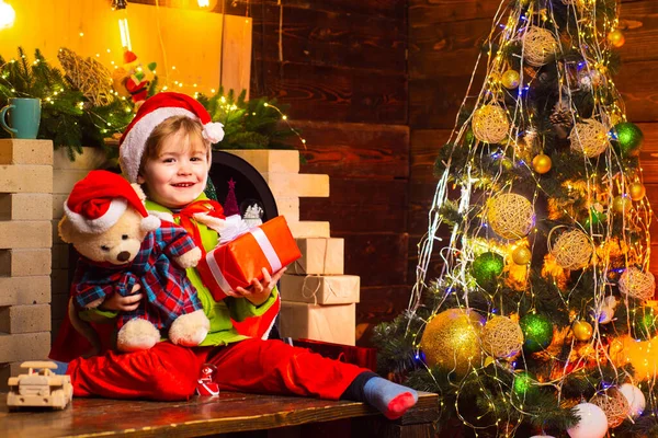 Alegre bebé sonriente Santa Boy sosteniendo su oso de peluche y regalo de Navidad cerca de la chimenea y árbol de Año Nuevo decorado. Niño pequeño decorando árbol de Navidad y regalos de apertura. —  Fotos de Stock