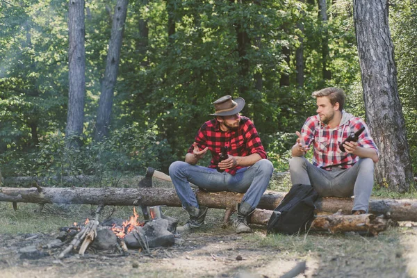 Gesellschaft erwachsene Freunde, die sich am Lagerfeuer entspannen. Gruppe von zwei männlichen Rucksacktouristen, die sich am Lagerfeuer entspannen. zwei befreundete Holzfäller sitzen im Wald und trinken Bier. — Stockfoto