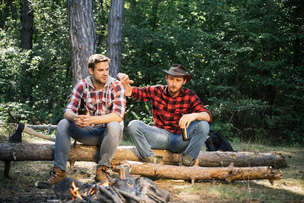 Young people enjoying picnic in park on summer day and drinking beer. Tourism concept. Picnic friends. Two friends lumberjack worker sitting in the forest and drinking beer.