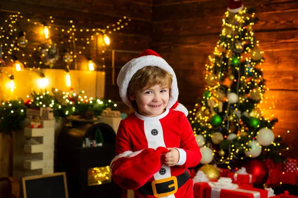 Weihnachtsgeschenke online kaufen. Weihnachtseinkaufskonzept. Urlaub und Winterkindschaftskonzept. Lächelnder kleiner Junge mit Weihnachtsmann-Helfermütze über dem Weihnachtsbaum zu Hause. — Stockfoto