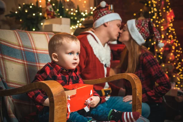 かわいい赤ちゃんがクリスマスのギフトボックスを開けています。背景にロマンチックな焦点を当てていない親とキス。大晦日の夜。奇跡です。ギフトカードの概念。クリスマスの属性。クリスマスツリー. — ストック写真