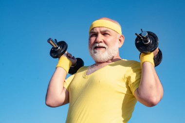 Bearded Senior sportman exercising with lifting dumbbell on blue sky background. Isolated, copy space. Senior man workout in rehabilitation center. Like sports.