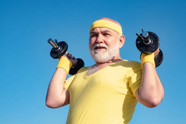 Bearded Senior sportman exercising with lifting dumbbell on blue sky background. Isolated, copy space. Senior man workout in rehabilitation center. Like sports. — 스톡 사진