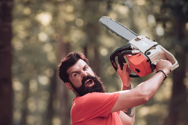 Excited lumberjack holding chainsaw in the forest. Woodcutter with chainsaw on sawmill. Handsome young man with axe near forest. — 스톡 사진