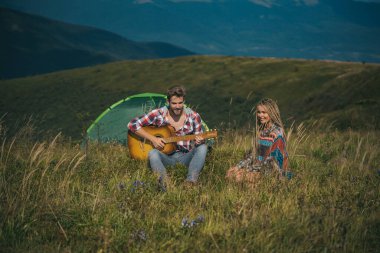 Kampın dışında, dağların tepesinde kamp kuran harika genç bir çift. Mutlu gezginler dinleniyor ve gitar çalıyor. Aktif yaşam tarzı ve inanılmaz dağ konsepti.