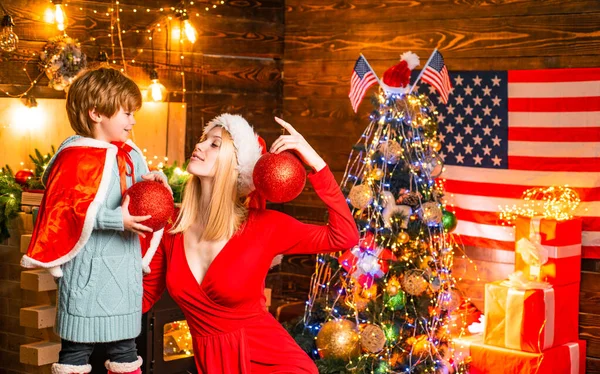 Hermosa madre joven en vestido rojo celebrar la Navidad con su querido hijo y decorar la habitación. Atributos de Navidad. Recuerdos de la infancia. Feliz niño pequeño jugar con decoraciones de Navidad . —  Fotos de Stock