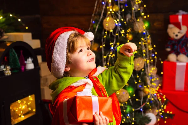 Niño feliz con regalo de Navidad está mirando hacia arriba. Niño feliz con regalo de Navidad. Retrato de Santa niña con regalo mirando a la cámara. Niño divirtiéndose cerca del árbol de Navidad en el interior . — Foto de Stock