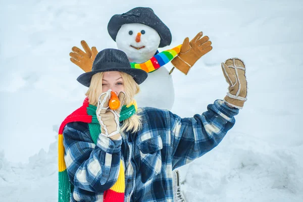 Fille drôle faisant bonhomme de neige avec nez de carotte drôle. Bonhomme de neige drôle avec une carotte au lieu d'un nez et dans un chapeau tricoté chaud sur une prairie enneigée sur un fond de neige floue . — Photo