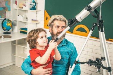 Baba oğluna öğretiyor. Komik yüzlü sevimli bir öğrenci. Astronomi teleskopu tutuyor..