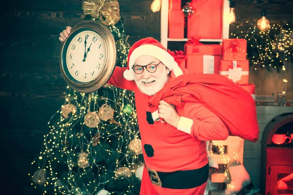 Fröhlicher Weihnachtsmann mit Wecker am Kamin und Weihnachtsbaumhintergrund. Der Weihnachtsmann zu Hause. Weihnachtsmann hält Wecker vor Weihnachtsbaum-Hintergrund. — Stockfoto